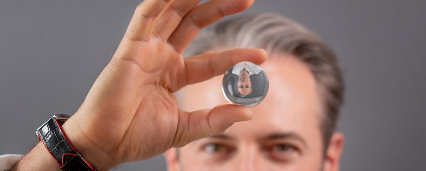 Jan Bruch holding a glass lens up to the camera; his face appears inverted inside it, himself soft-focus behind.