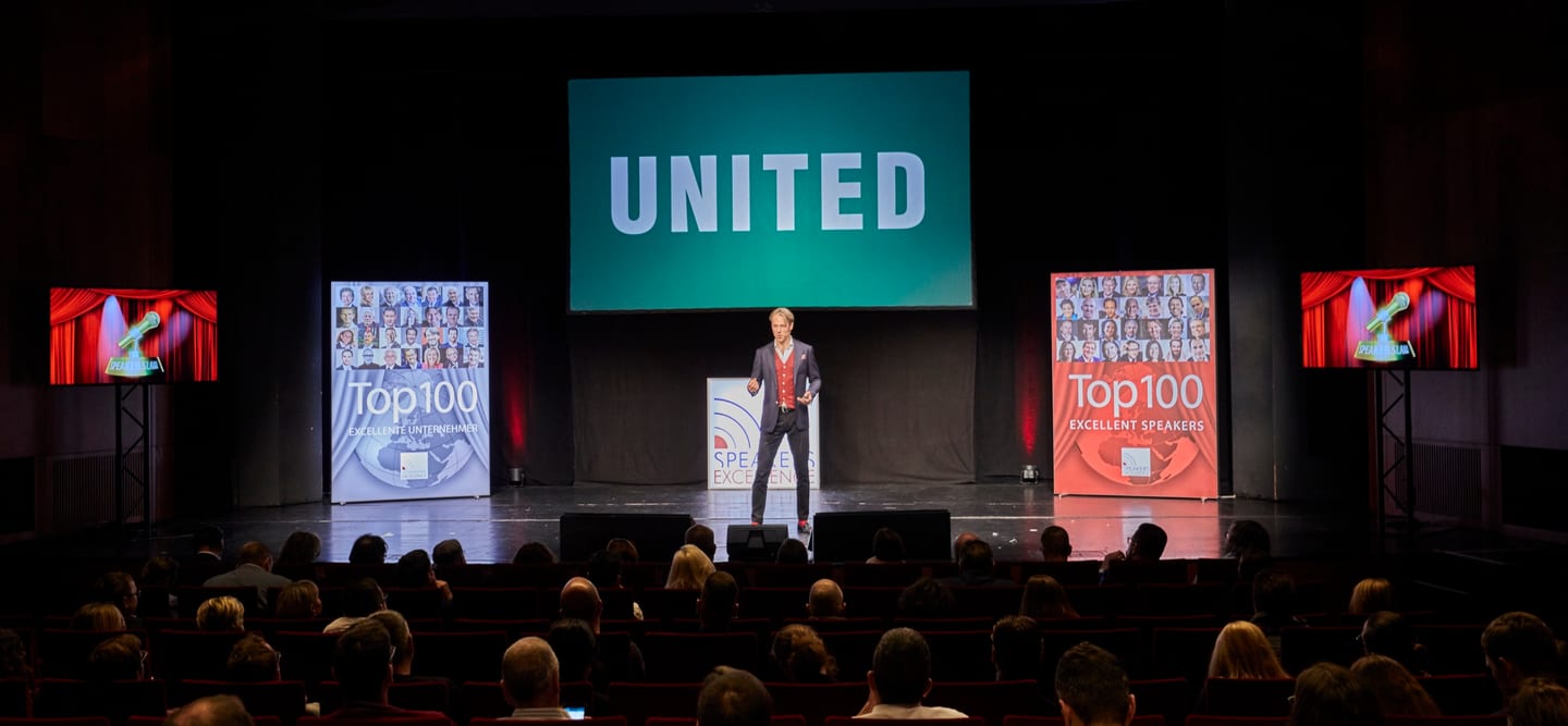 Jan Bruch alone on a large stage before a full auditorium, the word UNITED projected behind him.