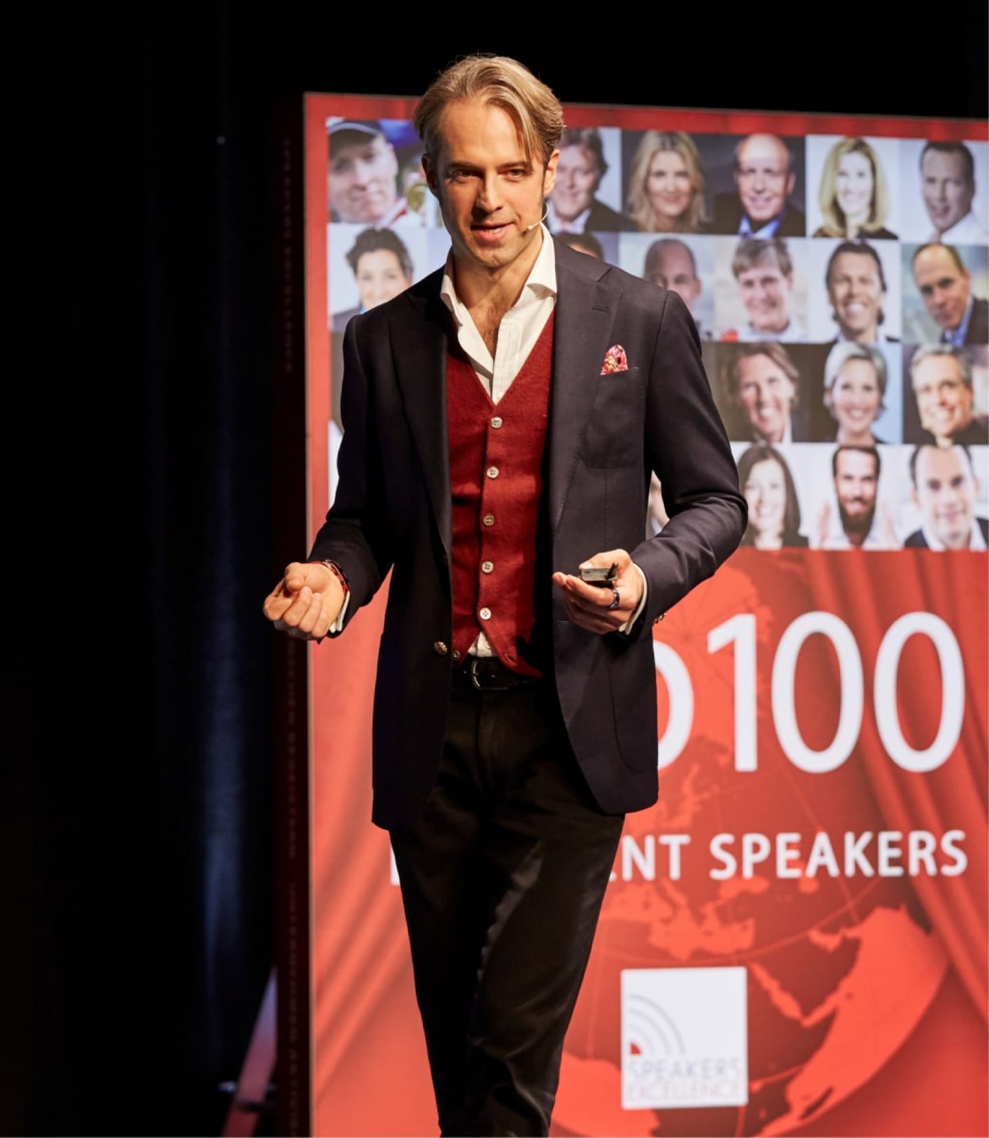 Jan Bruch walking a stage with a clicker, beside a Top 100 Excellent Speakers banner.
